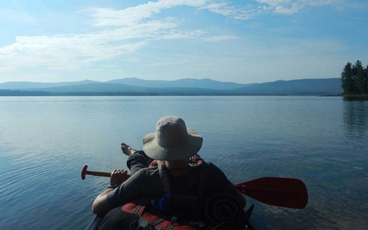 From the back of a canoe, you can see the person in front relaxing with their paddle. The water is calm and blue, and there are mountains in the distance.